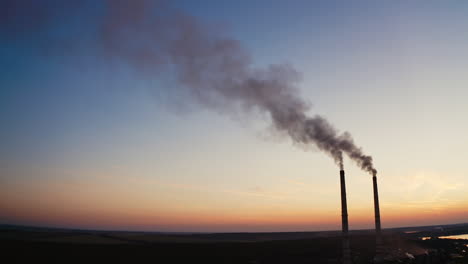 Factory with pipes among nature in the evening. Two industrial pipes with dark smoke pollute the environment at sunset.
