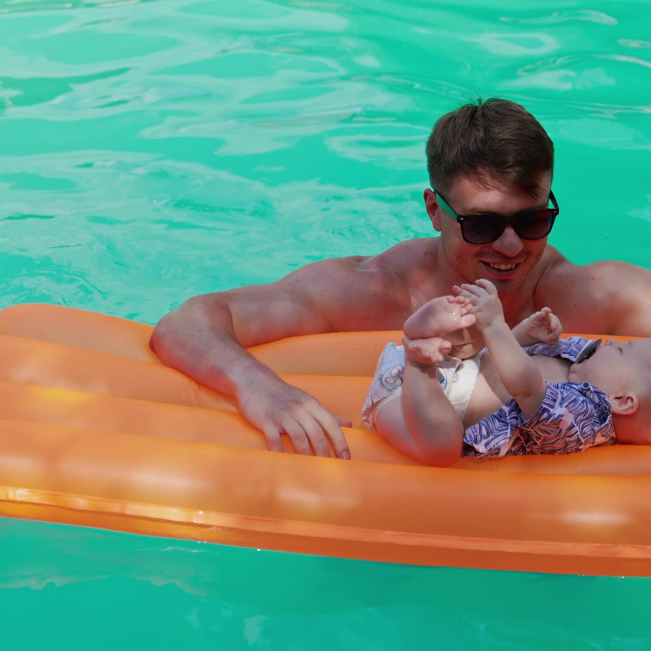 Caucasian baby in diaper and shirt with pacifier in mouth lies on the inflatable mattress. Father is standing at the mattress in the pool and kid cries. Vertical screen