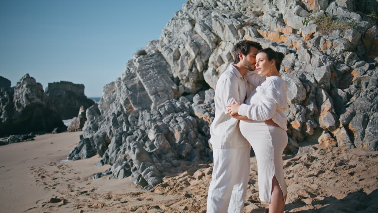 hombre cariñoso abrazando a una mujer embarazada de pie en una playa rocosa soleada. futuros padres