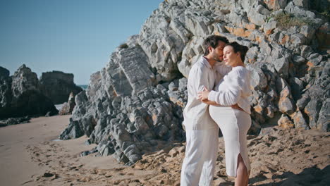 caring man embracing pregnant woman standing sunny rocky beach. future parents