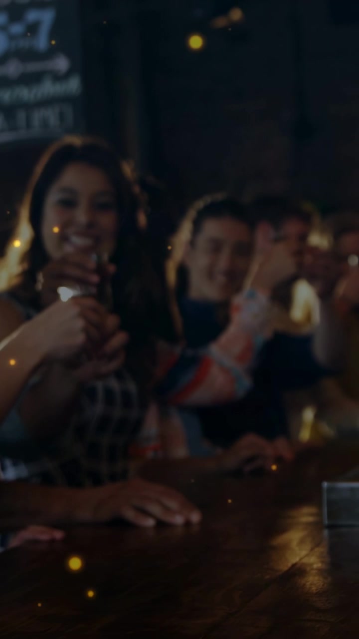 Video verticale: Donna che alza un bicchierino, dando il via a un brindisi in un bar, panoramica che mostra una cassetta per le mance, sovrapposizione dorata.