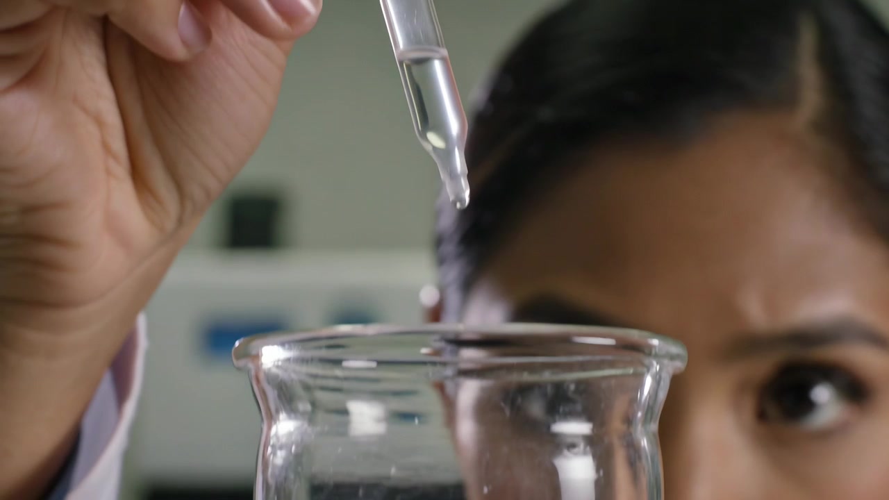 Usando un contagocce, scienziato indiano in camice da laboratorio che rilascia colorante blu in un becher sul banco, osservando la miscelazione.