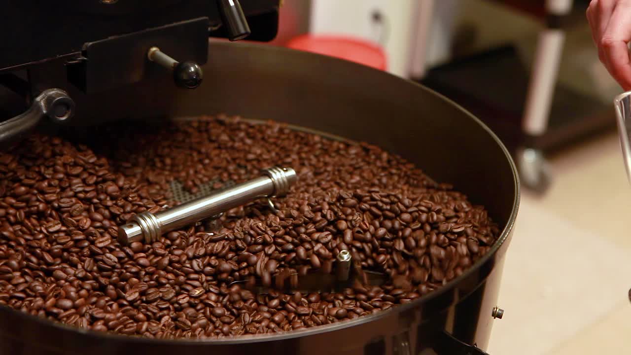 Person's hand uses a small clear scoop to push freshly roasted coffee beans as they are moved and mixed in roaster's cooling tray.