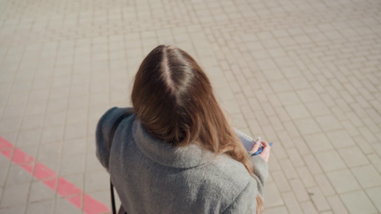 Young woman walking with notebook and phone, viewed from behind on tiled plaza, gray coat and black bag, overhead sunlight casting geometric shadows, quiet stride and minimalist urban routine