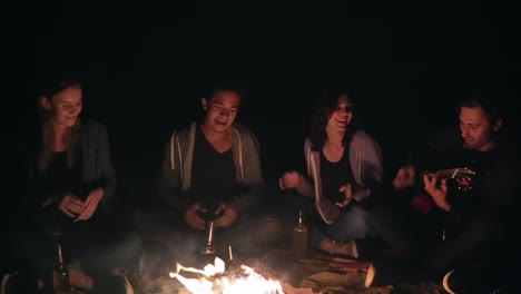 Closeup-view-of-the-bonfire-late-at-night.-Young-people-sitting-by-the-fire-in-the-evening,-playing-guitar.-Cheerful-friends-singing-songs,-talking-and-having-fun-together