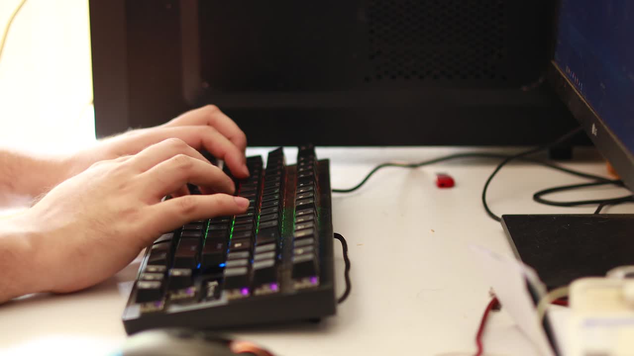 toma en ángulo alto desde el lado de las manos de un hombre interactuando con un teclado rgb en un escritorio de computadora sucio