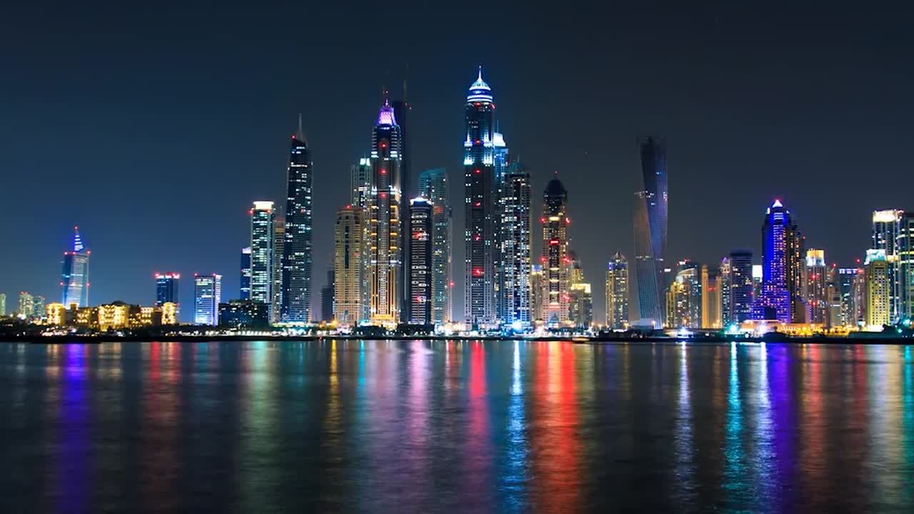 Filmato in time-lapse dello skyline illuminato di Dubai di notte, con grattacieli colorati che si riflettono splendidamente sull'acqua calma in primo piano. La scena evidenzia l'architettura moderna, vibrante.