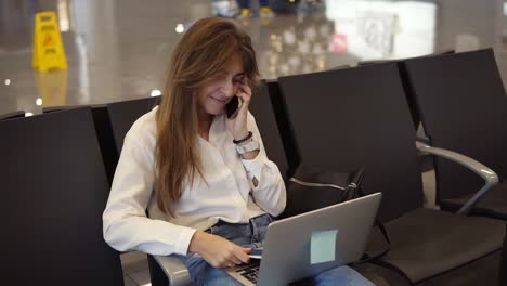 Stylisches,-Hübsches-Mädchen-Im-Weißen-Hemd-Benutzt-Telefon-Und-Laptop,-Um-Am-Flughafen-Zu-Arbeiten,-Während-Es-In-Der-Abflughalle-Auf-Das-Boarding-Wartet
