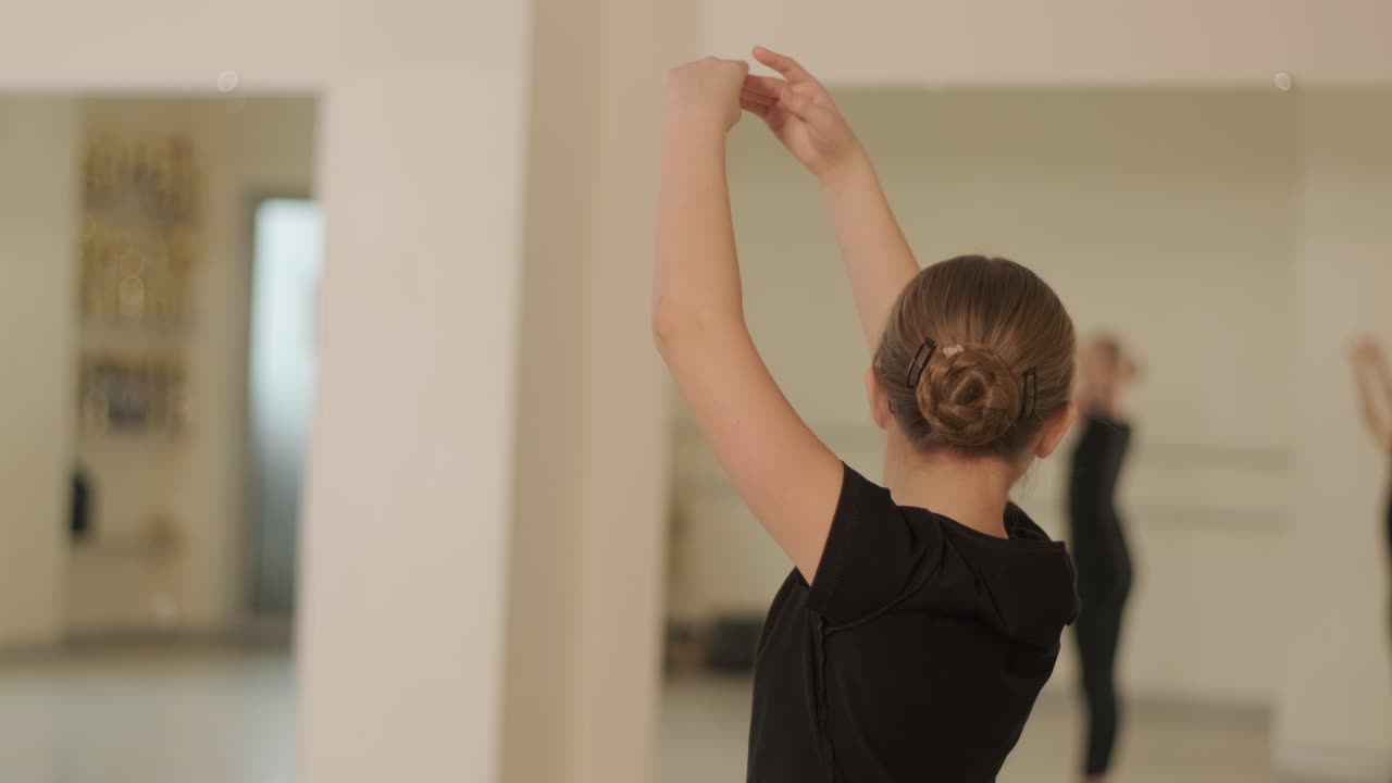 En gruppe unge ballettelever i svart dansebekledning som øver på posisjoner i et romslig ballettstudio med tregulv og veggmonterte barrer. Fokuserte uttrykk og synkroniserte bevegelser