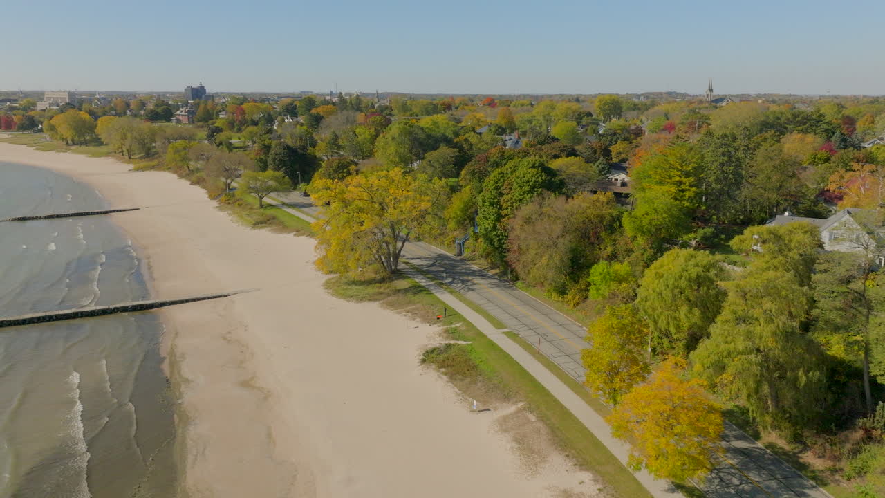 Drone aerial along the Lake Michigan shoreline and road toward a Sheboygan, Wisconsin neighborhood with houses and colorful autumn trees on a clear fall day