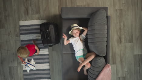 toddler walks past sister resting on sofa and big suitcase