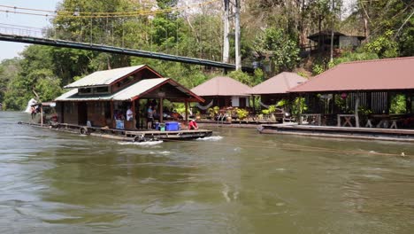 a very charming wooden floating house is sailing under a bridge and passing a small floating village in the middle of the jungle of sai yok national park in thailand in asia on a clear blue day