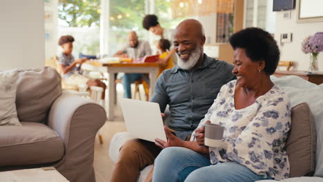 Senior-Couple-At-Home-Looking-At-Laptop-Together-With-Multi-Generation-Family-In-Background