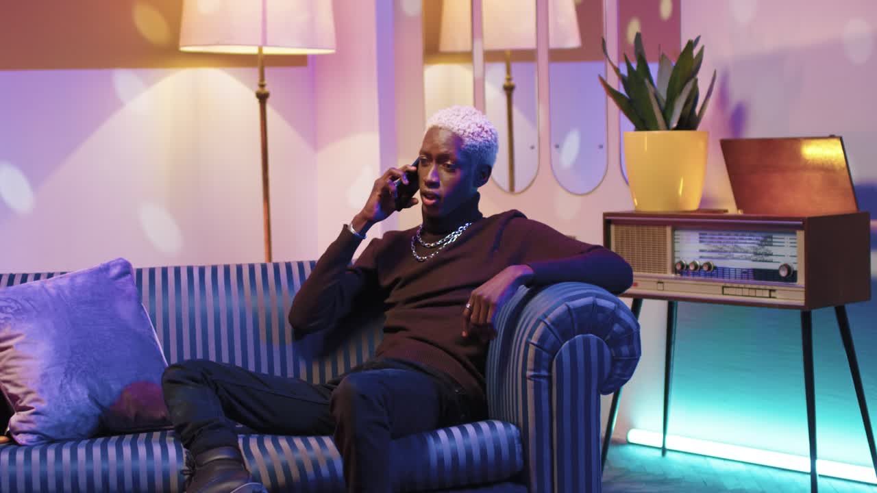 African american man with blonde hair talking on the smartphone while sitting on sofa in a retro room at home