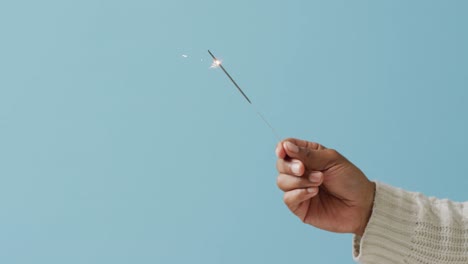 Video-of-hand-of-african-american-person-holding-lit-sparkler-on-blue-background,-with-copy-space