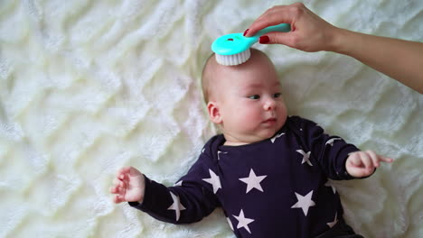 healthy caucasian toddler lies on bed and moves actively. mother's hand with a brush touches baby head and he smiles with pleasure. top view.