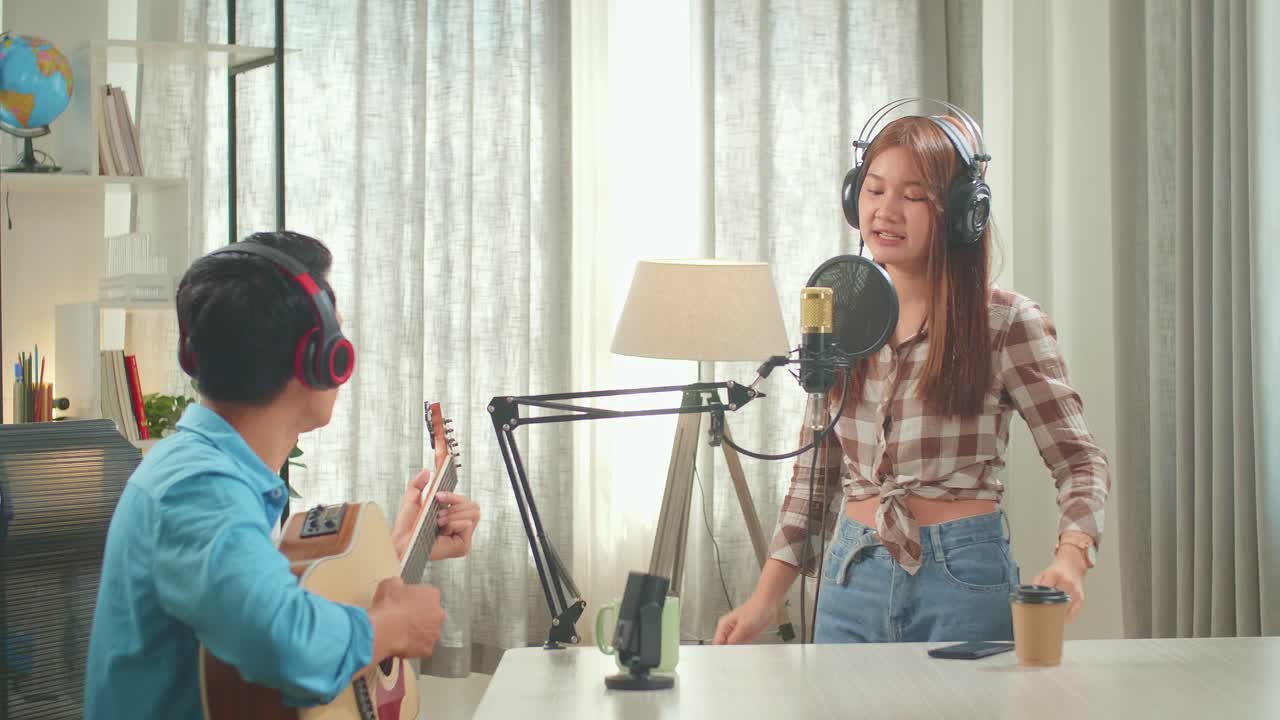 Young Asian Woman In Headphones Singing Into Microphone And While Man Playing The Guitar During Online Livestream In Podcast Studio