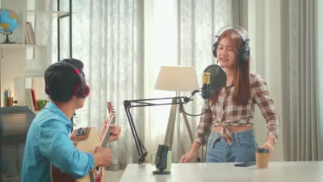 young asian woman in headphones singing into microphone and while man playing the guitar during online livestream in podcast studio