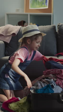 funny little girl with straw hat puts garment heap and checks up belongings in open case at home. smart kid gathers luggage for distant travel