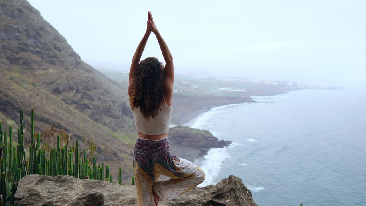 Practicing yoga in island mountains, a young woman stands on one leg, arms extended, enjoying ocean views