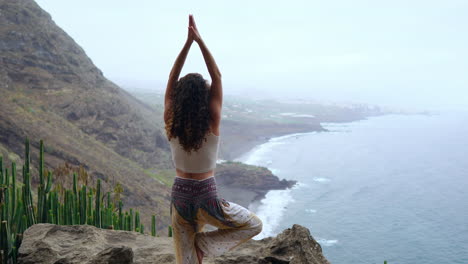 practicando yoga en las montañas de la isla, una mujer joven está de pie sobre una pierna, brazos extendidos, disfrutando de las vistas del océano