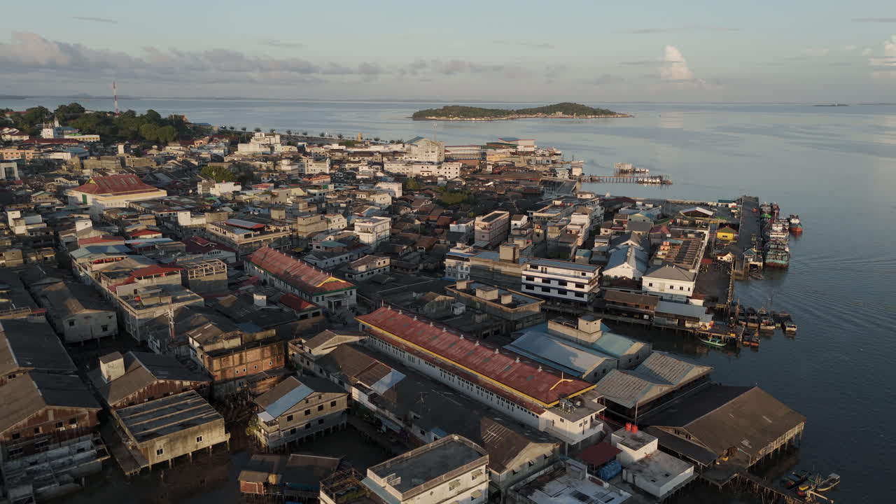 Gentle aerial view of the bustling downtown port and dense coastal city of Tanjung Pinang at sunrise. Buildings built over the water, small docks, and a soft golden light. Tanjungpinang, Indonesia