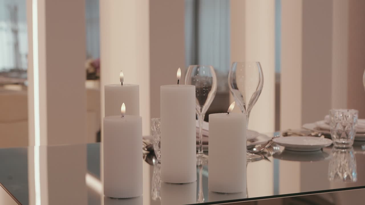 Candele bianche alte che brillano su un tavolo riflettente vicino a bicchieri e piatti.