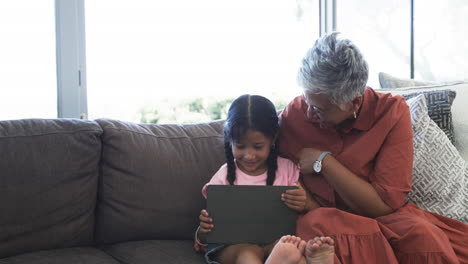 Una-Chica-Birracial-Con-Una-Tableta-Se-Sienta-Al-Lado-De-Su-Abuela,-Compartiendo-Un-Momento-De-Alegría-Con-Espacio-Para-Copiar