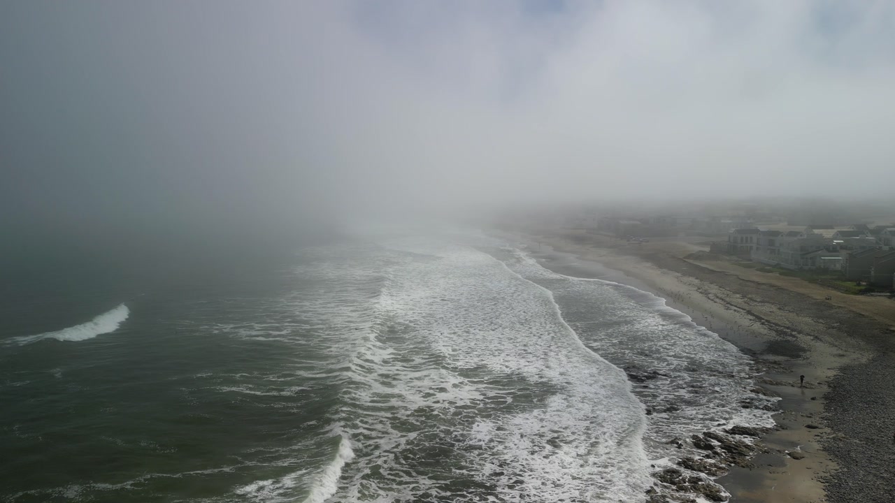 Drone video capturing the foggy Cape Town coastline, with mist over waves breaking on the sandy beach. Highlights the dramatic seaside under overcast skies, offering a moody coastal vista.