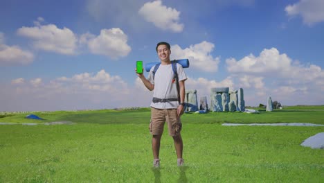 full body of asian male hiker with mountaineering backpack smiling and showing mock up green screen smartphone while traveling in stonehenge