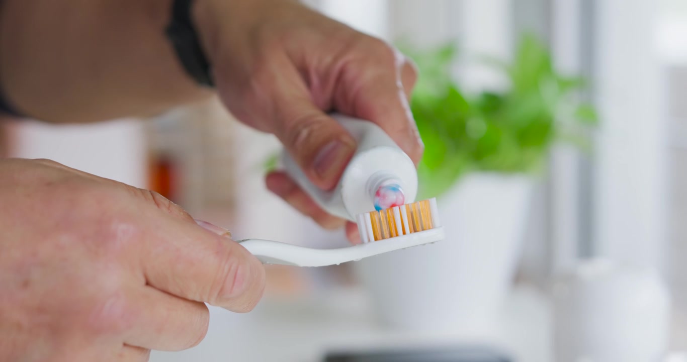 Senior Asian man holding toothbrush squeezing tube at sink, applying tricolor paste for brushing