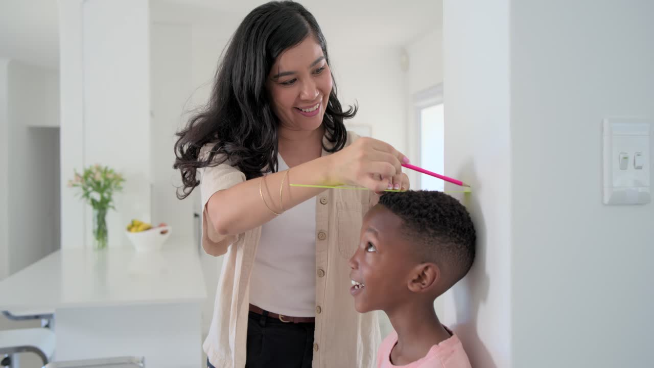 Diverse mother measuring son on white wall with yellow strip, pink marker tracking growth