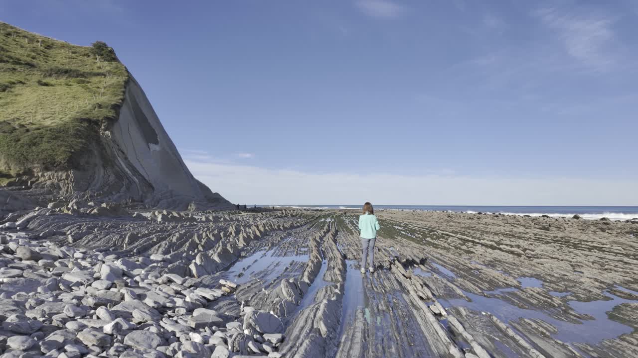 Giovane donna avventurosa che cammina verso l'oceano sull'impressionante Via del Flysch, una formazione geologica unica sulla costa dei Paesi Baschi tra Zumaia e Deba in una giornata di sole.