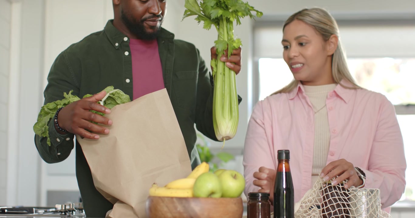 Una coppia afroamericana disfa la spesa sull'isola della cucina, l'uomo mette la mano nella borsa selezionando verdure per i pasti.
