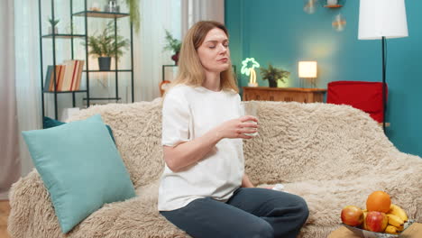 Pregnant woman swallows tablet drinks water sighs relief vitamin treatment routine on sofa at home