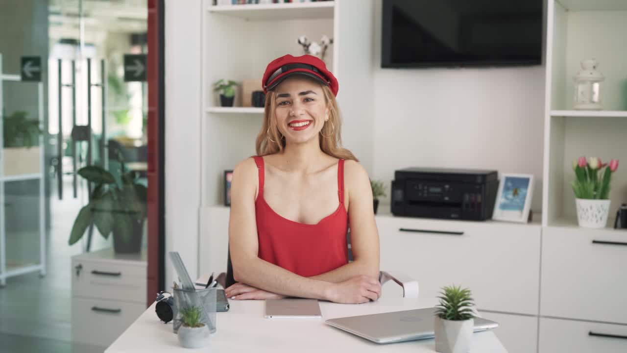 The young woman in a red dress and black hat is sitting at a table in the office and smiling at the camera. There is a portrait of a young woman in the office