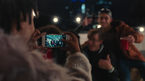 young happy friends posing for photo on rooftop at night enjoying party african american woman photographing reunion gathering using smartphone sharing on social media