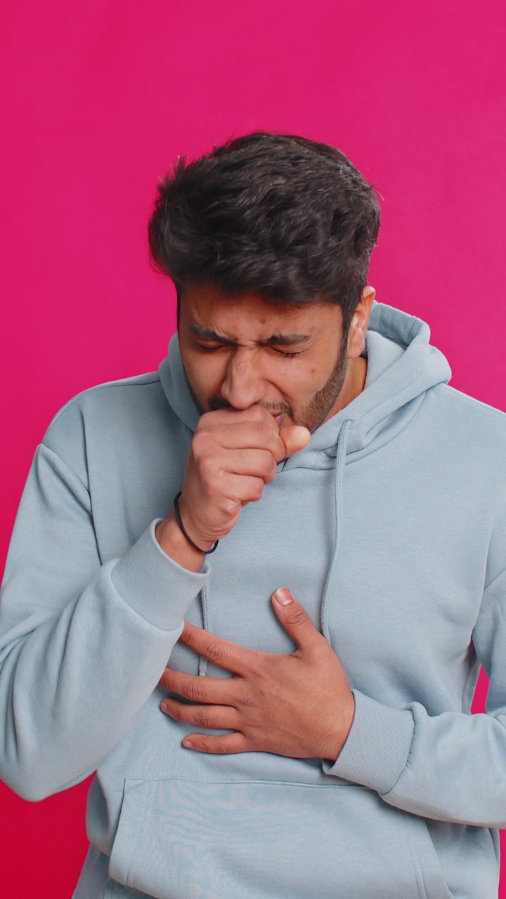 Joven indio enfermo tosiendo, cubriéndose la boca con la mano, sintiéndose mal sobre un fondo rosa.