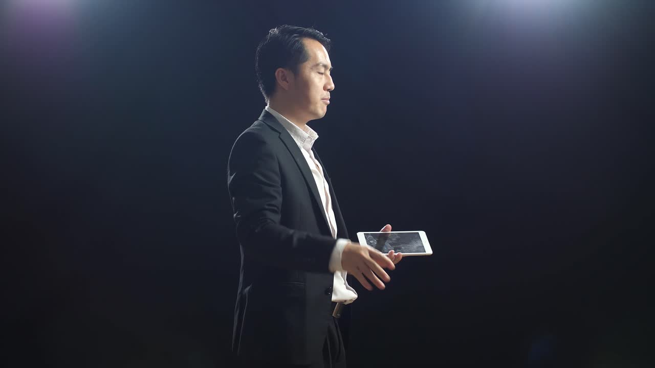 블랙 스크린 스튜디오에서 말하는 동안 태블릿을 들고 가리키는 비즈니스 슈트를 입은 아시아 스피커의 측면 시각