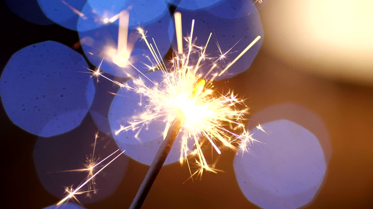 A close-up macro shot of burning New Year sparkler. Bengal light scattering into small sparkles against a background of blue garlands in blur. 4k slow motion