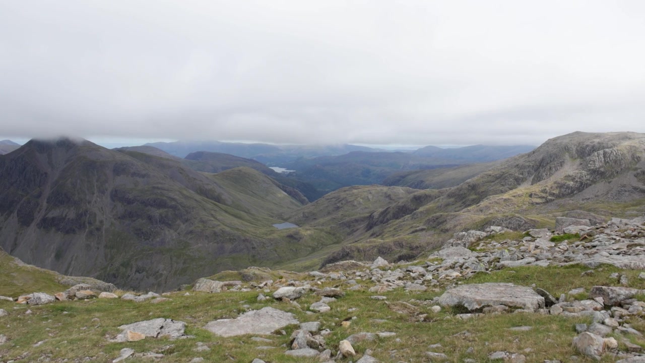 Colline meridionali da Skafell Pike, panoramica verso destra attraverso le aspre creste montuose del Lake District, primo piano con cima rocciosa, strato di nuvole, valli e laghi alpini distanti in Cumbria, Inghilterra