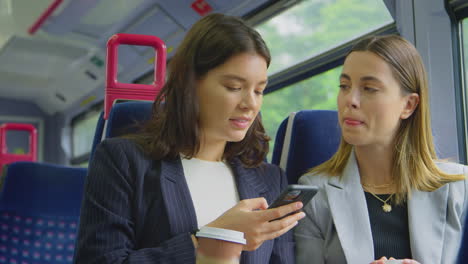 Two-Businesswomen-With-Takeaway-Coffees-Commuting-To-Work-On-Train-Looking-At-Mobile-Phone-Together