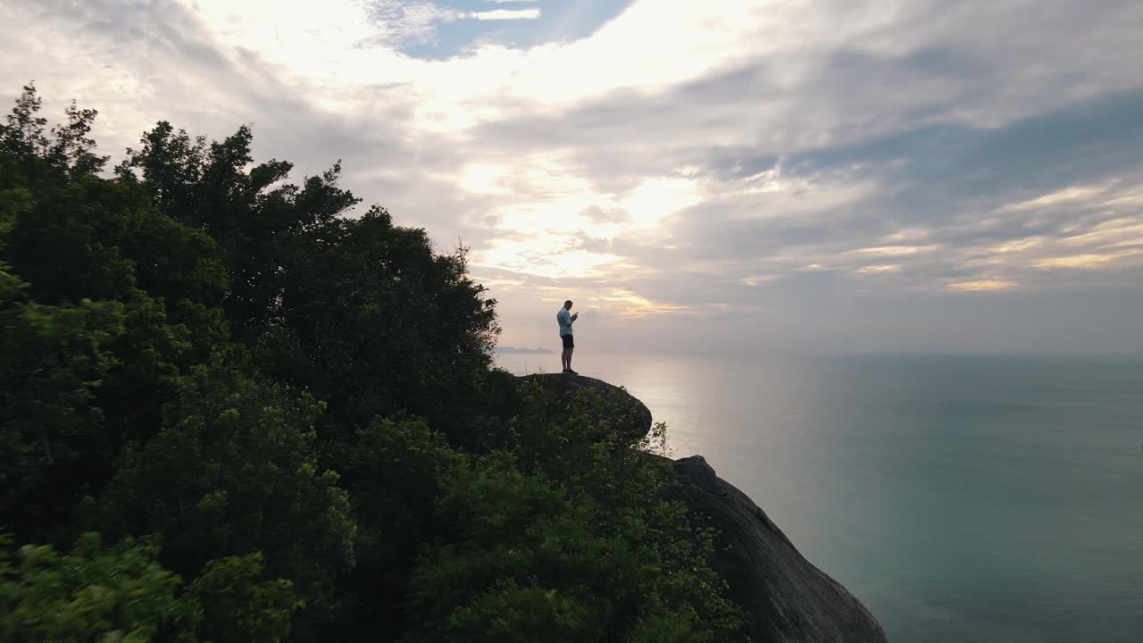 hombre europeo se encuentra en una gran roca entre los árboles que se mueven salvajemente en el viento con una hermosa vista sobre el mar claro en koh pha ngan en un día nublado
