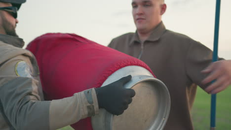 close up of two people handling hot air balloon basket as man removes gas cylinder covered in red cloth during preparation on open field near parked vehicle