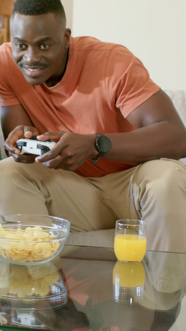 Vertical video: Reacting to challenge, gamer in rust tee gripping gamepad at home on sofa smiling