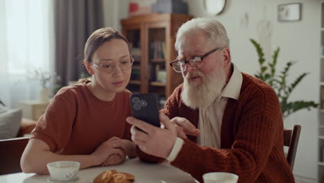 young woman teaching senior grandfather how to use modern smartphone at home