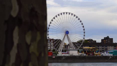 Zeitlupenaufnahme-Von-Einem-Baumstamm-Zu-Einem-Sich-Bewegenden-Riesenrad-Bei-Einem-Volksfest-In-Köln-Am-Rhein-Mit-Anderen-Beleuchteten-Fahrgeschäften-Während-Der-Abenddämmerung