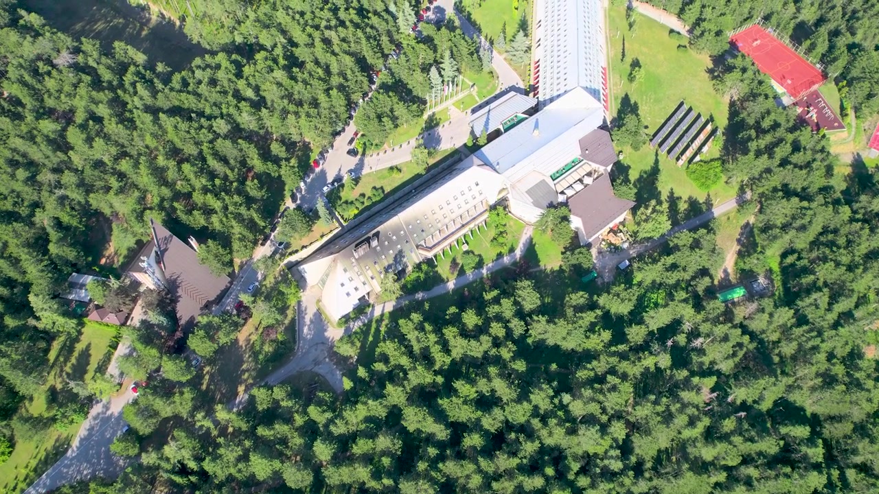 A top-down aerial drone approaches Hotel Omorika on Tara Mountain through dense green canopy, revealing the white hotel, lawns, red-roof building and paved grounds in daylight.