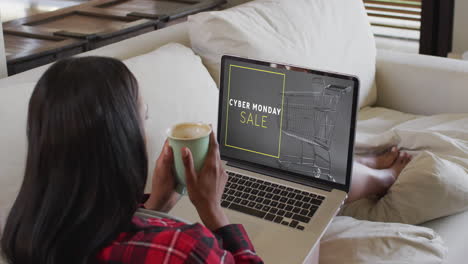 biracial woman on couch with coffee using laptop for online shopping, slow motion