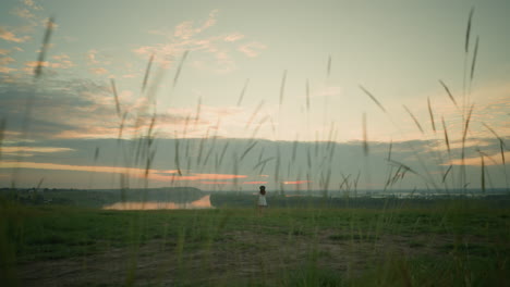 a woman in a white dress and hat stands alone in an isolated grassy field by a tranquil lake at sunset. she gazes at the serene surroundings, embracing the peaceful and reflective moment